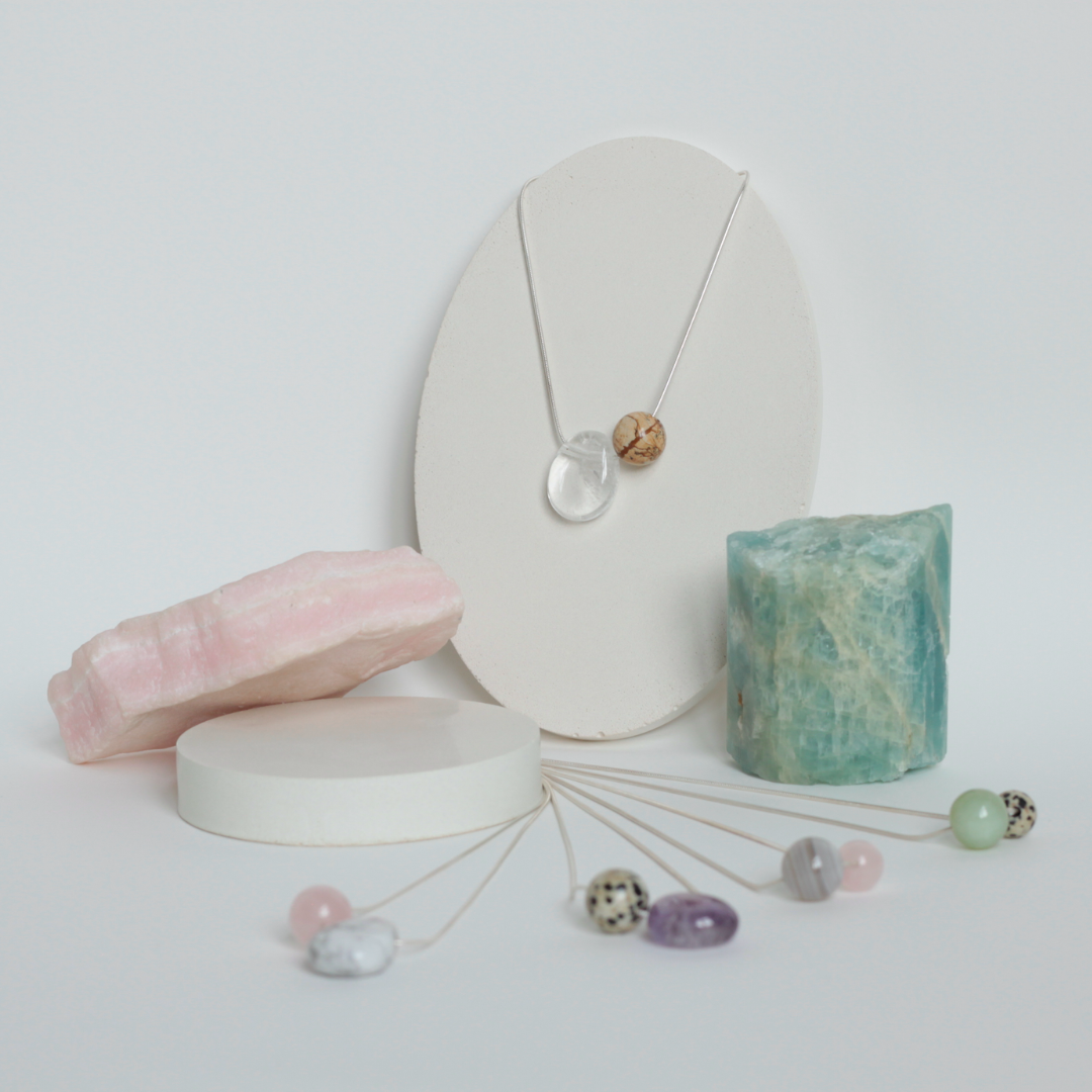 Asymmetrical Semiprecious Gemstone Crystal Pendant Necklaces with Clear Quartz, Beige and Brown Kalahari Jasper, Pink Rosequartz, Beige and Black Dalmatian Jasper, Lilac Amethyst, Grey and White Botswana Agate, Green Serpentine Asymmetrical Pendants and Round Beads on a Solid Sterling Silver Snake Chain with a Sterling Silver Clasp and Extension Chain in a Styled Studio Environment on a Jewellery Holder with Raw Gemstones as Props
