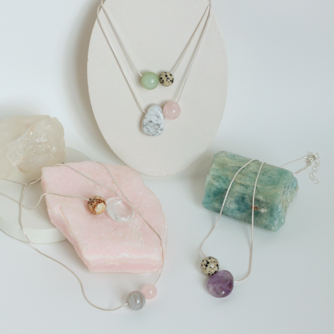 Asymmetrical Semiprecious Gemstone Crystal Pendant Necklaces with Clear Quartz, Beige and Brown Kalahari Jasper, Pink Rosequartz, Beige and Black Dalmatian Jasper, Lilac Amethyst, Grey and White Botswana Agate, Green Serpentine Asymmetrical Pendants and Round Beads on a Solid Sterling Silver Snake Chain with a Sterling Silver Clasp and Extension Chain in a Styled Studio Environment on a Jewellery Holder with Raw Gemstones as Props