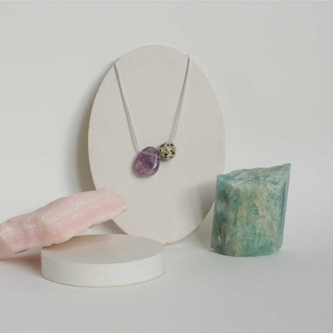 Asymmetrical Semiprecious Gemstone Crystal Pendant Neklace with an Asymmetrical Lilac Amethyst Pendant and Round Beige and Black Dalmatian Jasper Bead on a Solid Sterling Silver Snake Chain with a Sterling Silver Clasp and Extension Chain in a Styled Studio Environment on a Jewellery Holder with Raw Gemstones as Props